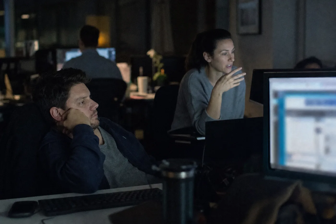 One worker slumped watching a glowing screen in the evening while a colleague leans in and speaks during a live interactive session