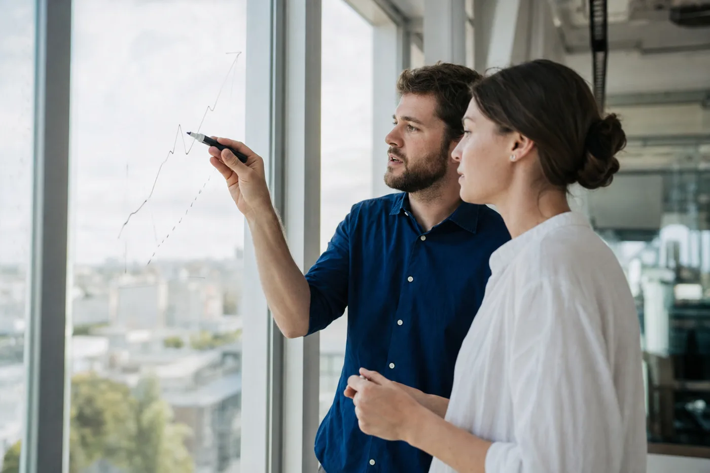 Two coworkers at a glass wall sketching a rising line by hand while planning a training program in bright daylight