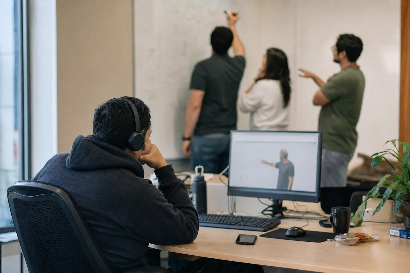 A passive learner watching a training video with headphones while colleagues behind him practice actively at a whiteboard