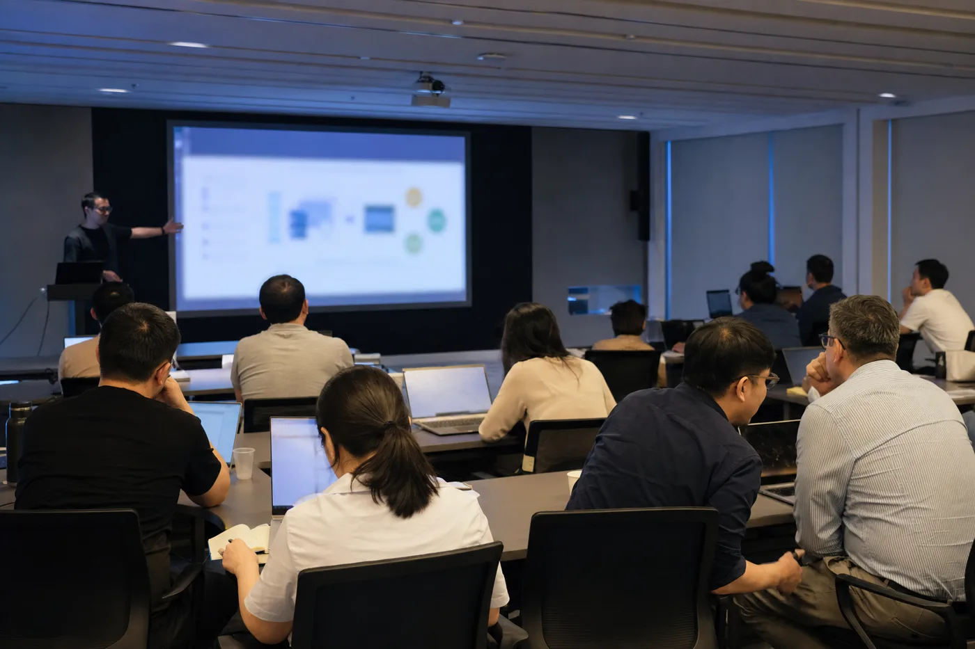 Employees in a company training room during an AI corporate training session, watching a presenter beside a projection screen