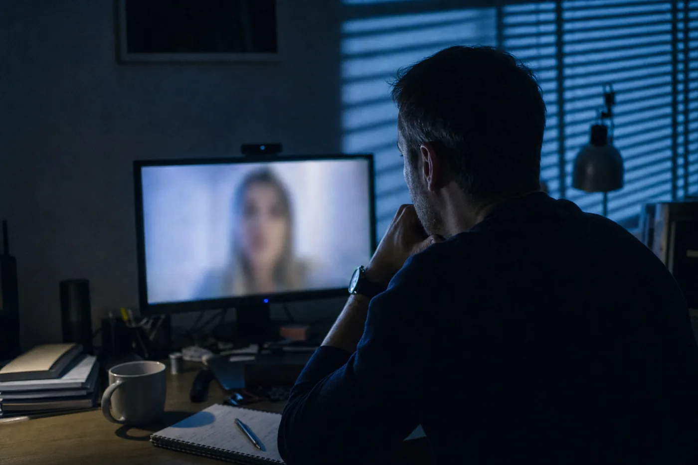 Over-the-shoulder view of a recruiter at a dim desk during an evening video interview, the person on the call soft-focus and unrecognizable, illustrating the deepfake candidate risk behind AI hiring fraud