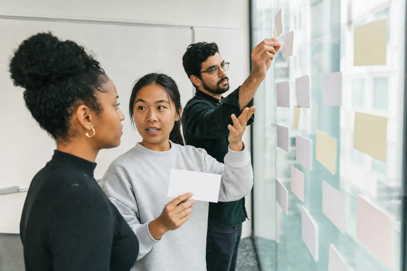 Three new employees doing a hands-on onboarding exercise together at a glass wall, arranging blank cards and sticky notes, representing interactive practice-based onboarding