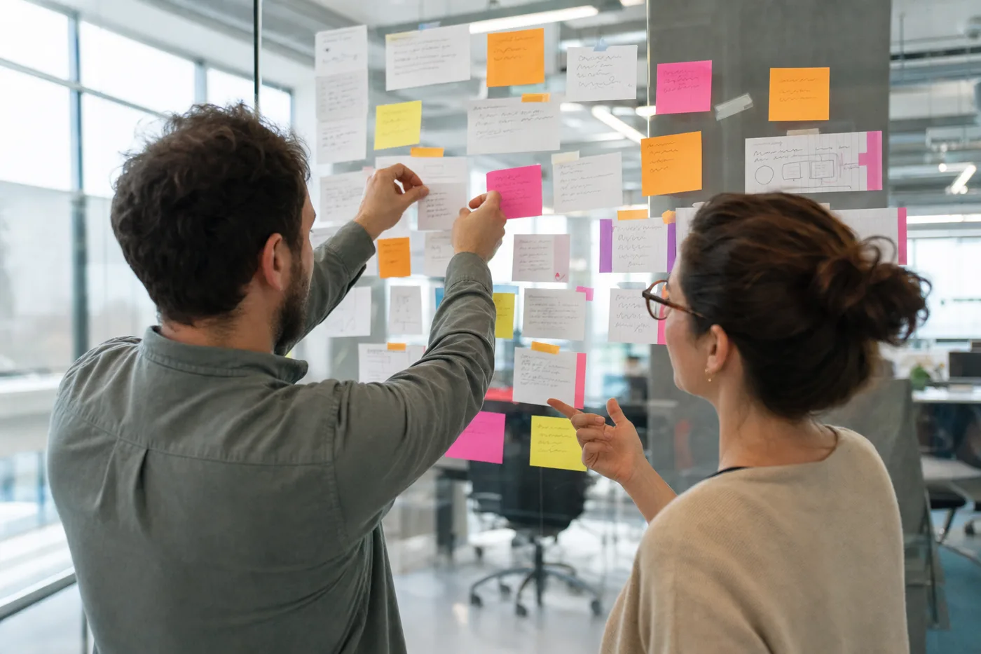 Hands arranging printed cards and colored sticky notes on a glass wall to map out a training curriculum, bright daylight office