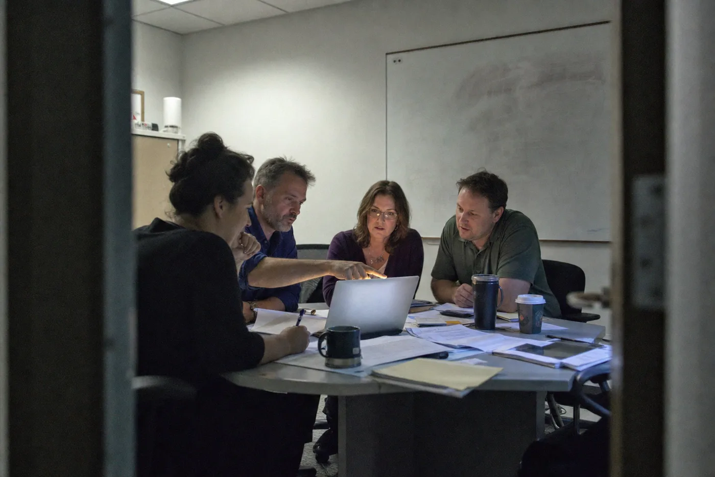 Four coworkers in a conference room reviewing assessment results together around a laptop and printed pages