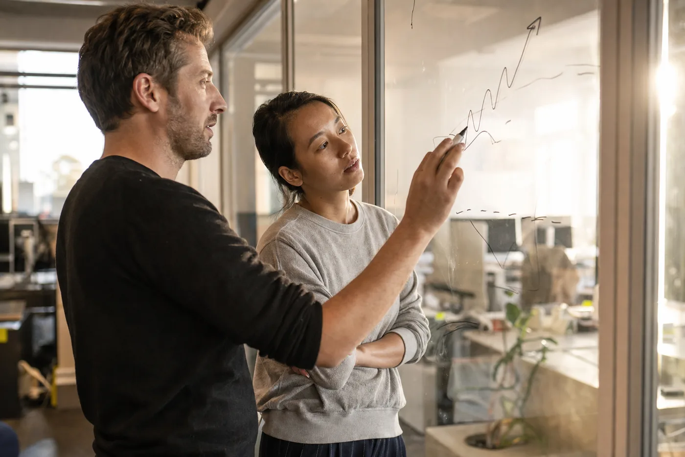 Two coworkers by a glass office wall discussing a rough rising curve sketched in marker, afternoon side light, candid and slightly grainy