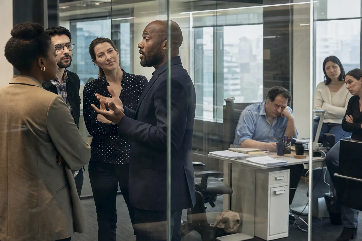A confident executive briefing a group by a glass wall on one side, and an uncertain manager with two employees at a desk on the other, in bright overcast daylight