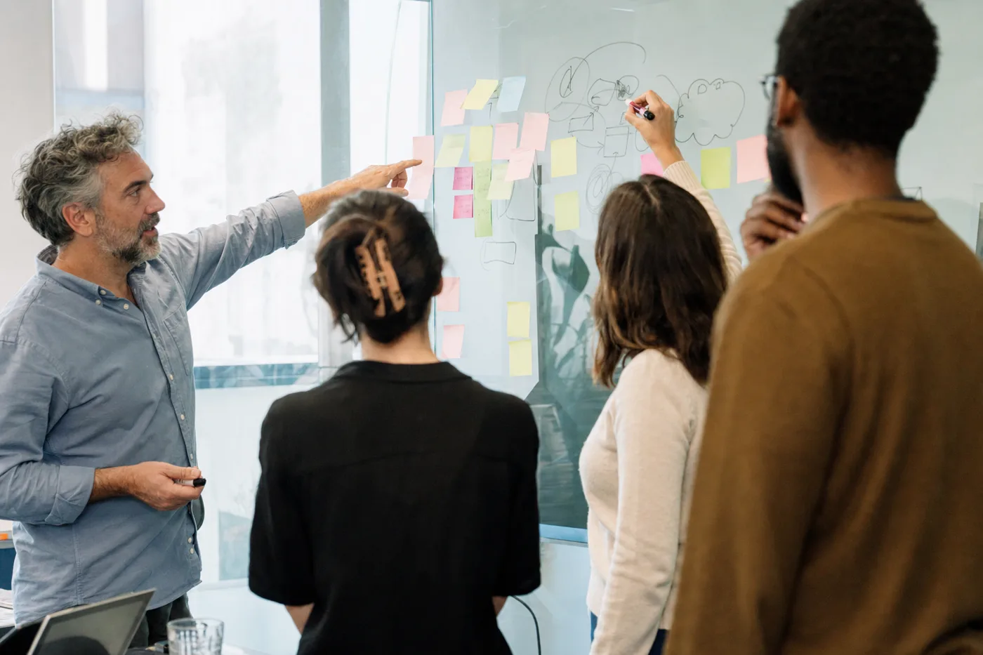 Four coworkers at a glass wall covered in sticky notes during a hands-on security awareness workshop, practicing how to respond to a suspected deepfake