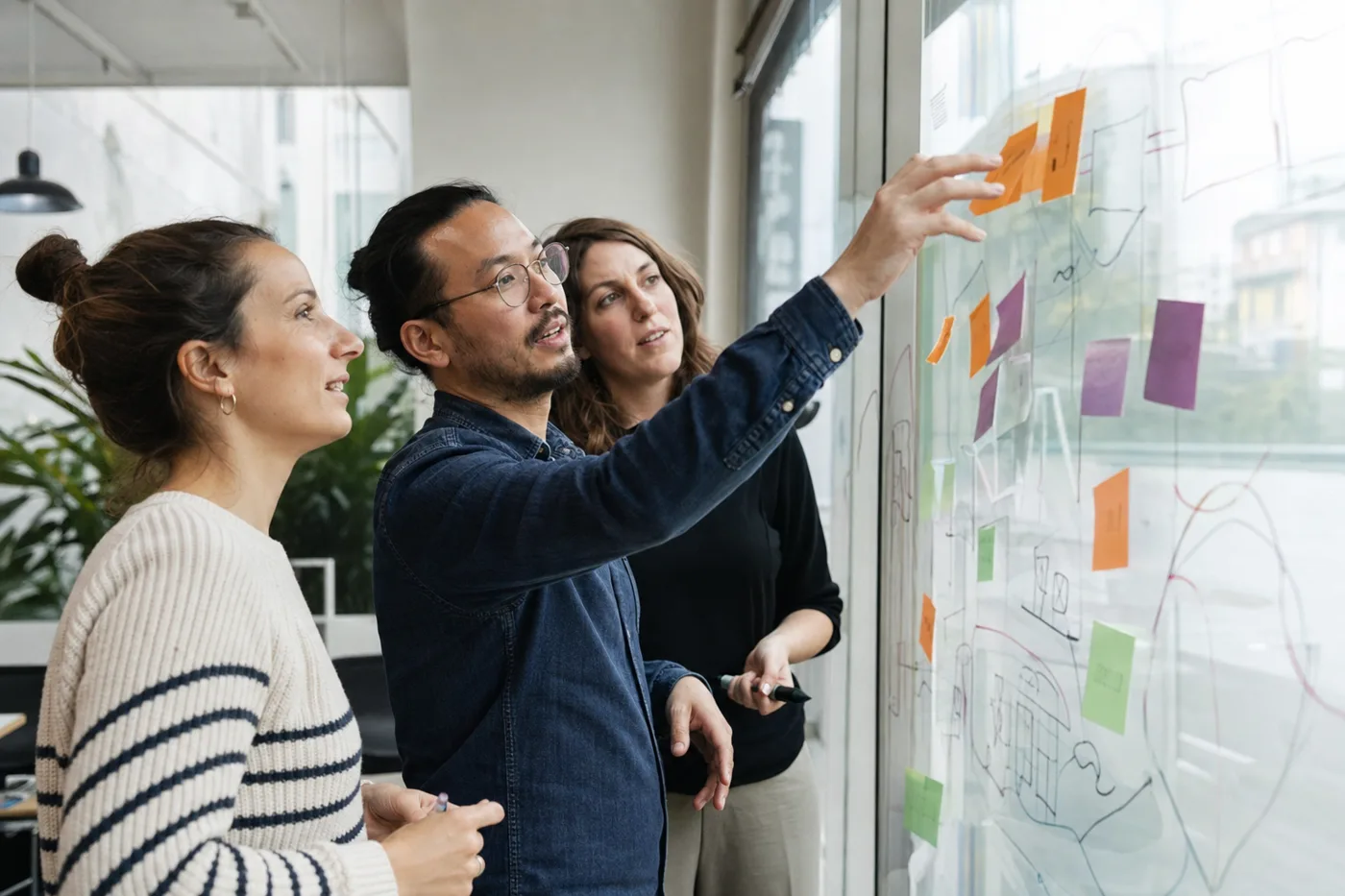 Coworkers standing at a glass wall arranging colorful sticky notes and hand-drawn diagrams during a collaborative workshop in bright daylight