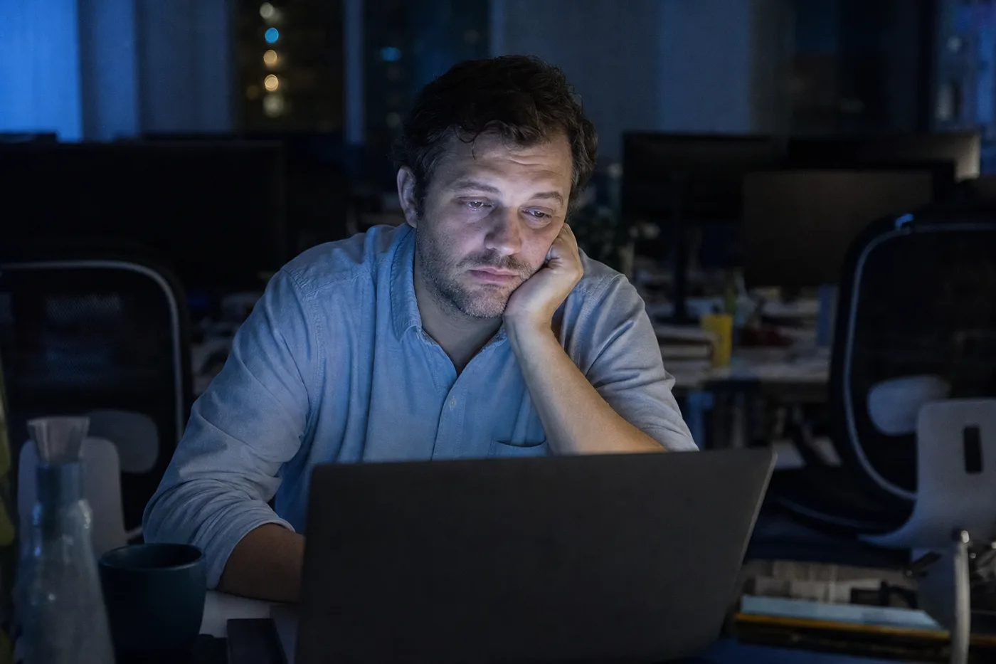 A tired employee half-watching a mandatory training video alone at a laptop in a dim office, the screen glow lighting their face