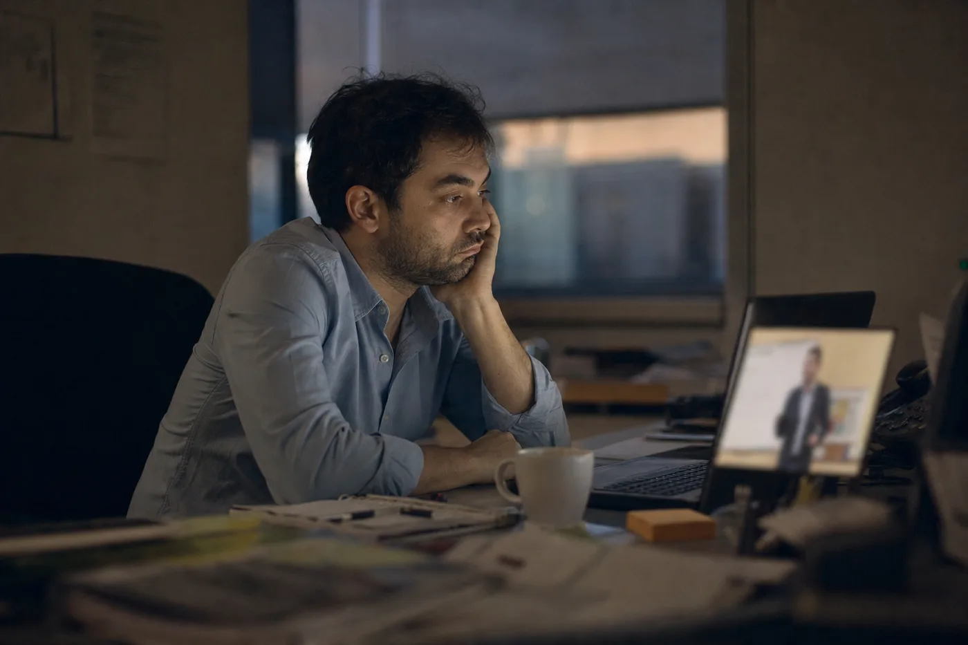 A lone employee half-watching a training video on a laptop in a dim office late in the afternoon, screen light on their face, looking disengaged