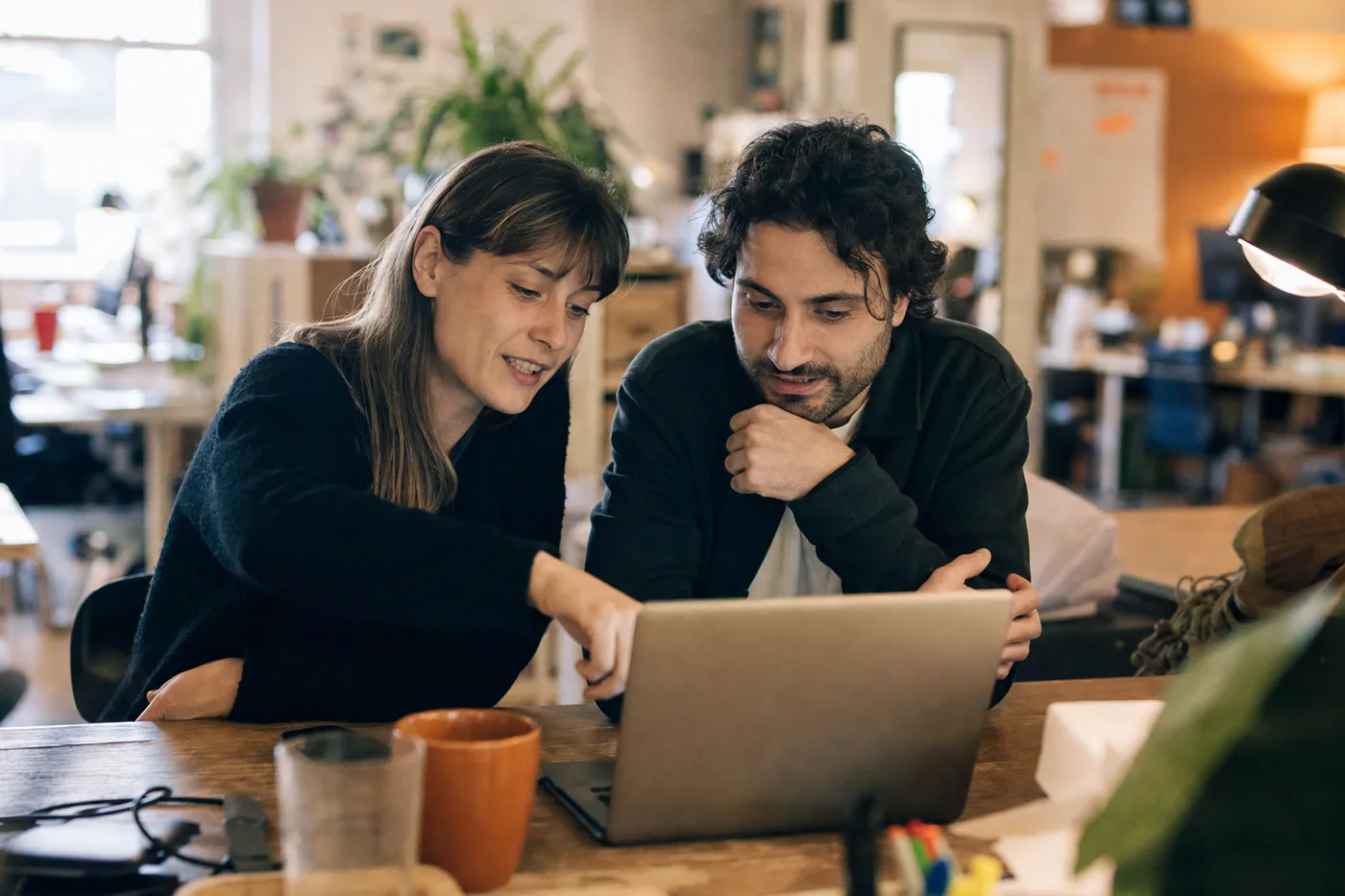 Two coworkers engaged in an interactive video training session at a shared desk, one leaning in and pointing at the laptop