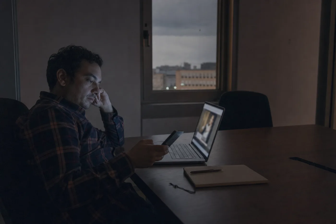 A tired employee half-watching a long training video alone in a dim meeting room, attention drifting to a phone