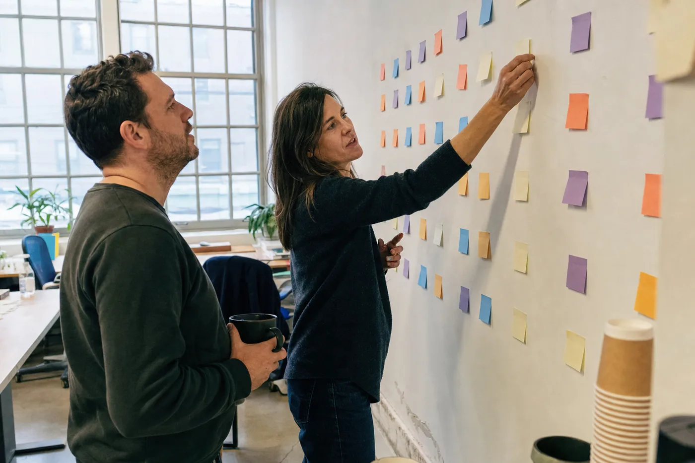 Two colleagues rearranging sticky notes on an office wall while mapping a course, the planning stage of learning engineering