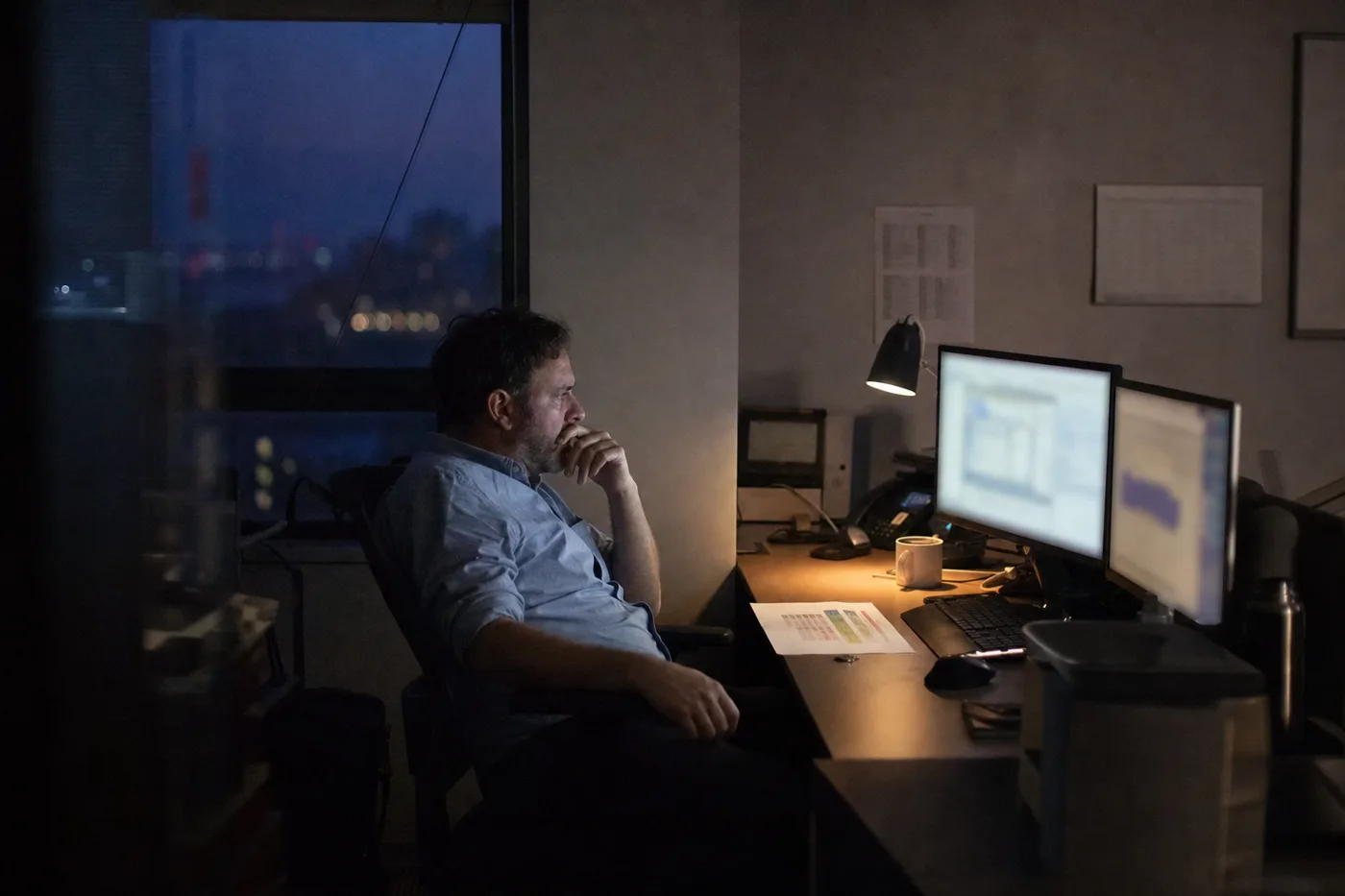 An analyst reviewing performance data on screen late in the evening, lit by monitor glow and a desk lamp