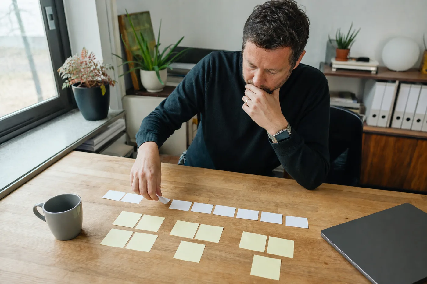 A man at a bright, window-lit desk pausing to arrange blank index cards and sticky notes as he checks his own understanding