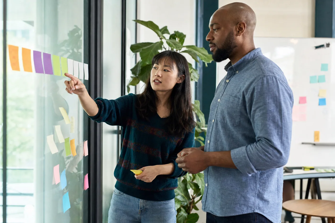 Two coworkers mapping a new hire's 30-60-90 day onboarding plan with sticky notes on a glass wall to speed time to productivity