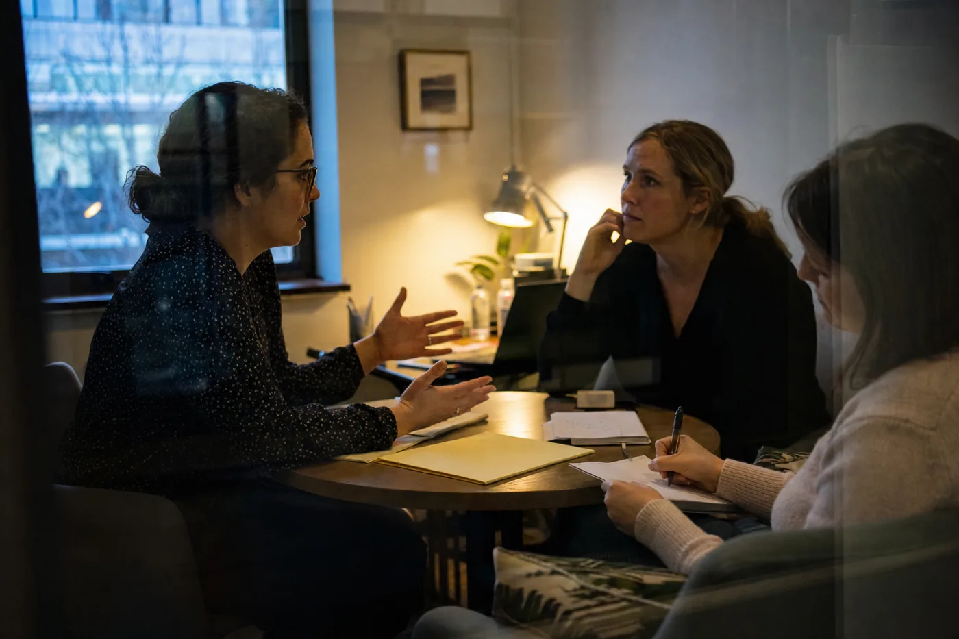 A woman practicing a client conversation with a colleague in a small meeting room while a coworker takes notes, candid documentary style
